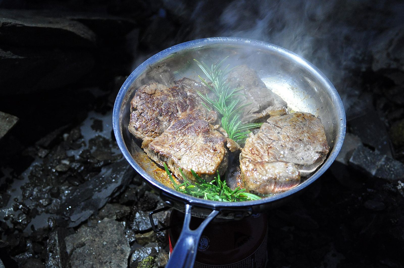 Food cooking in a pan on a camp stove set up outdoors at dusk.