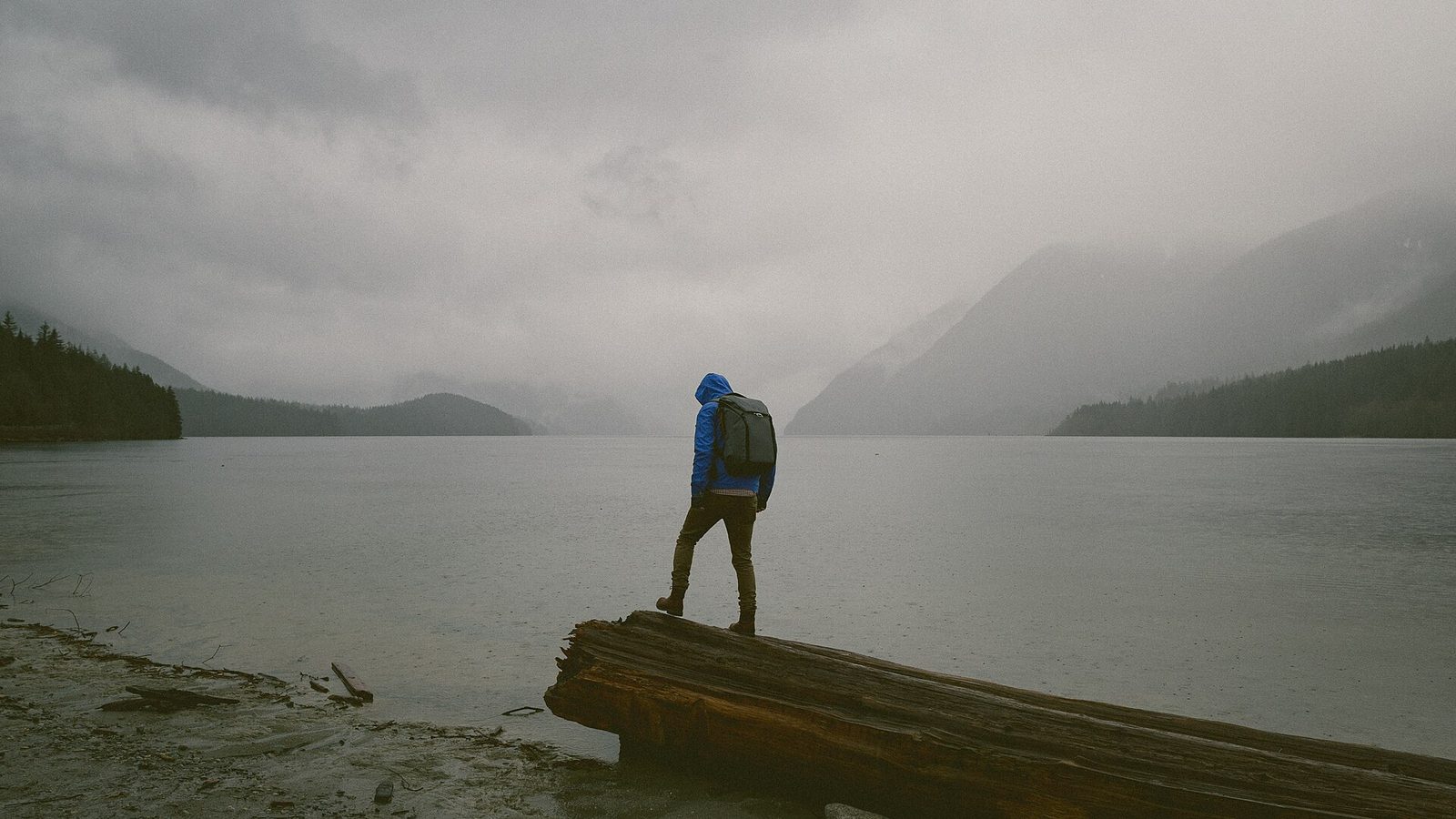 A hiker in a rain shell standing by a misty lake in steady drizzle.