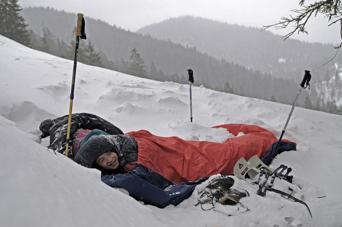 A person resting in a sleeping bag on snow with trekking poles beside them.